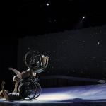 Laurel Lawson, a white person, balances above Alice Sheppard with arms spread wide, wheels spinning.  Alice, a multiracial Black woman with coffee-colored skin, opens her arms wide to receive her in an embrace. A starry sky fills the background, and moonlight glints off their wheelchair rims. Photo Jay Newman/BRITT.